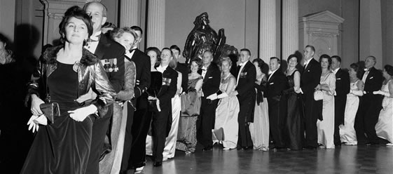 A monochrome image from 1964 depicting a line of people dancing while holding the waist of the person in front of them.